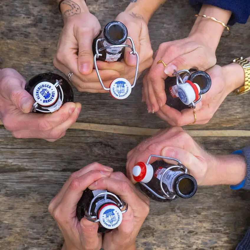 Vogelperspektive: Gruppe von Leuten stößt mit Bügelverschluss‑FLENS-Bierflaschen über rustikalem Holztisch an.