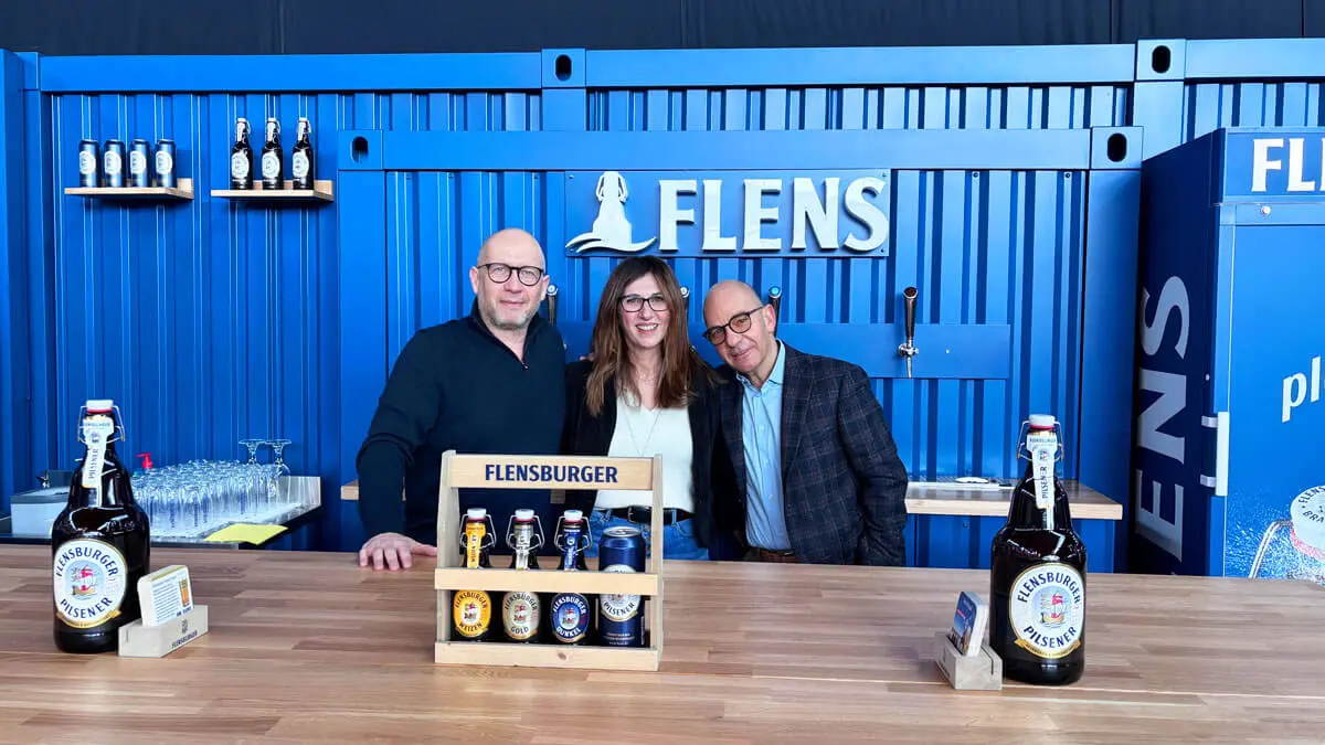 Flensburger Bier-Produkte vor der blauen Rückwand des Containermoduls des neuen Messestands, zwei Männer und eine Frau stehen hinterm Tresen.
