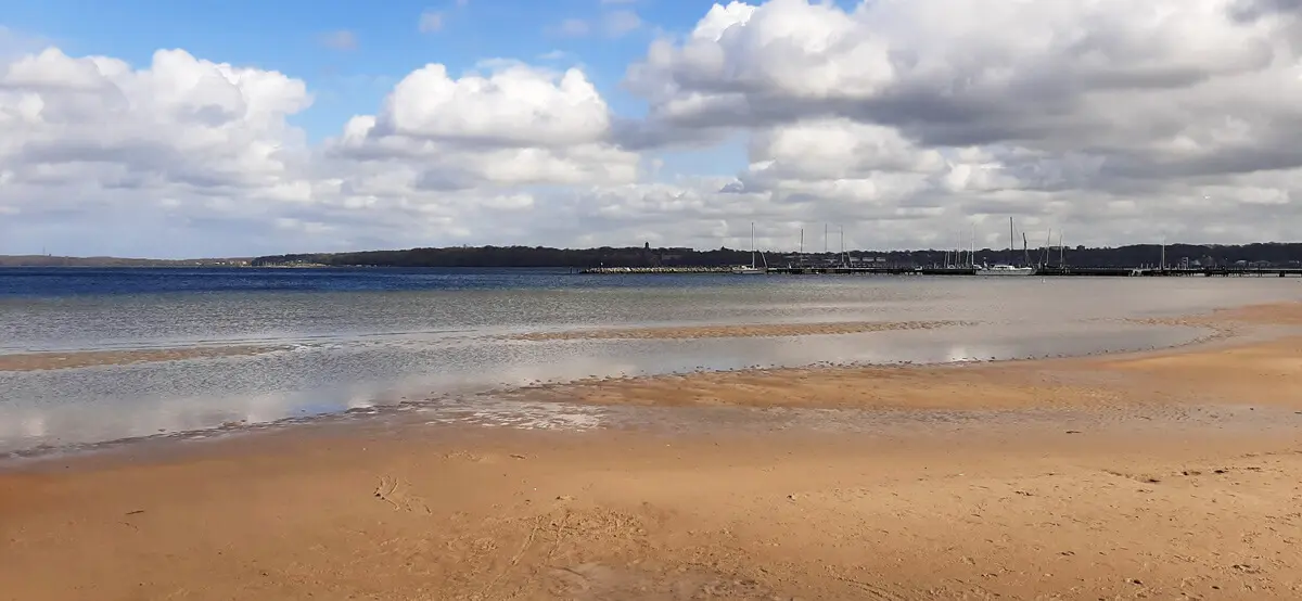 Sandstrand von Wassersleben in Flensburg mit flachem Wasser und Segelbooten im Hafen, ruhige Bucht unter bewölktem Himmel.