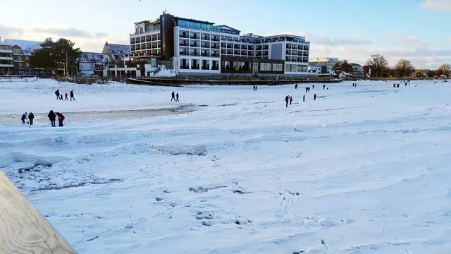 Vereister Strand mit Hotelpromenade, Spaziergängern und Fußspuren im Schnee.