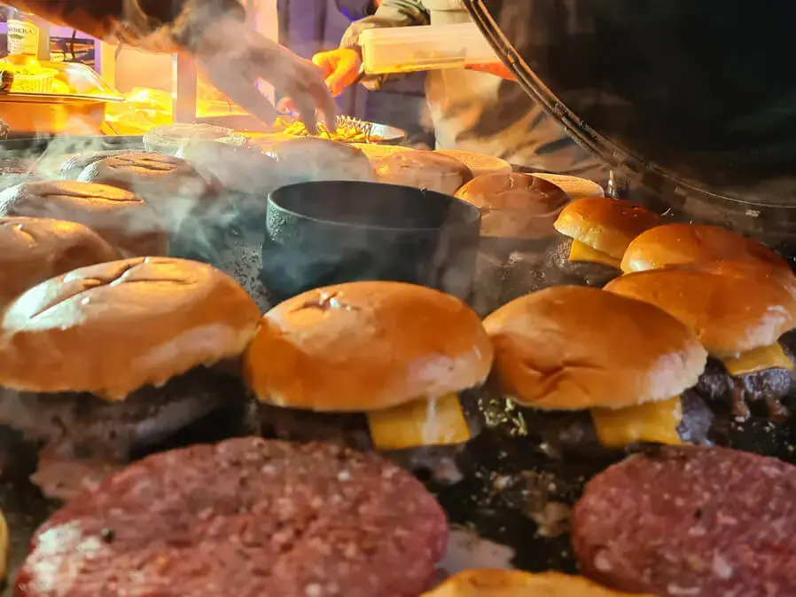 Burger werden auf einer Outdoor-Grillstation gebraten, Rauch und Dampf steigen auf.