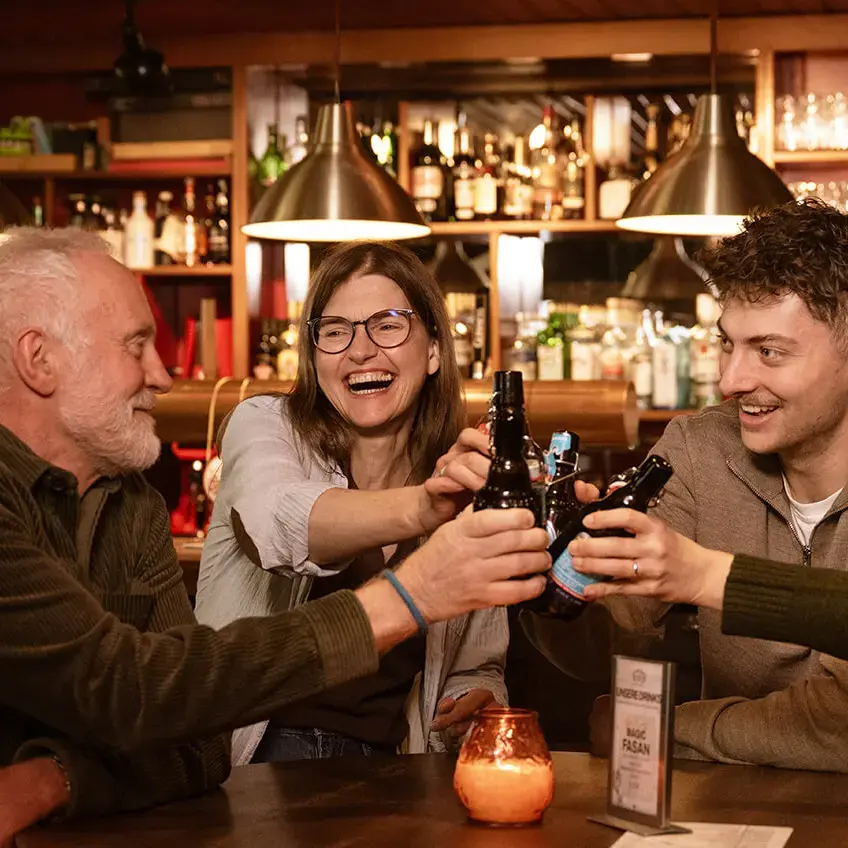 Vier Freunde stoßen mit FLENS Bierflaschen in gemütlicher Bar an, warmes Licht und Barregal im Hintergrund.