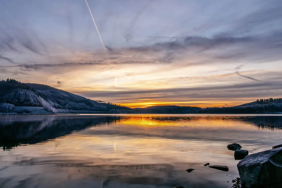 Sonnenuntergang über dem Biggesee mit Bergsilhouetten und goldener Spiegelung, Felsen im Vordergrund.