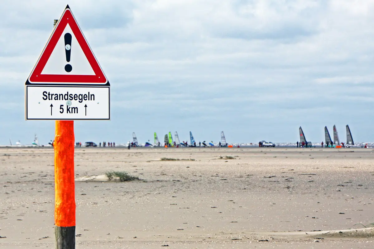 Warnschild "Strandsegeln 5 km" am Strand von Sankt Peter-Ording, Strandsegler und Landsegelboote am Horizont.