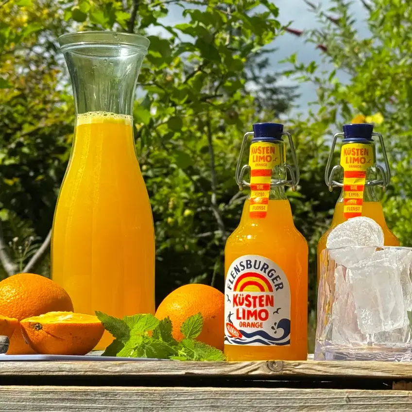 Eine Karaffe mit Orangelimonade, zwei Flaschen Flensburger Küstenlimo Orange, aufgeschnittene Zitrusfrüchte und ein Glas mit Eiswürfeln auf einem Tisch mitten im Grünen.