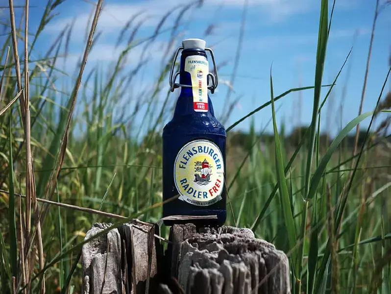 Eine blaue Flasche Flensburger Radler Frei auf einem Holzpoller im Schilfgras.