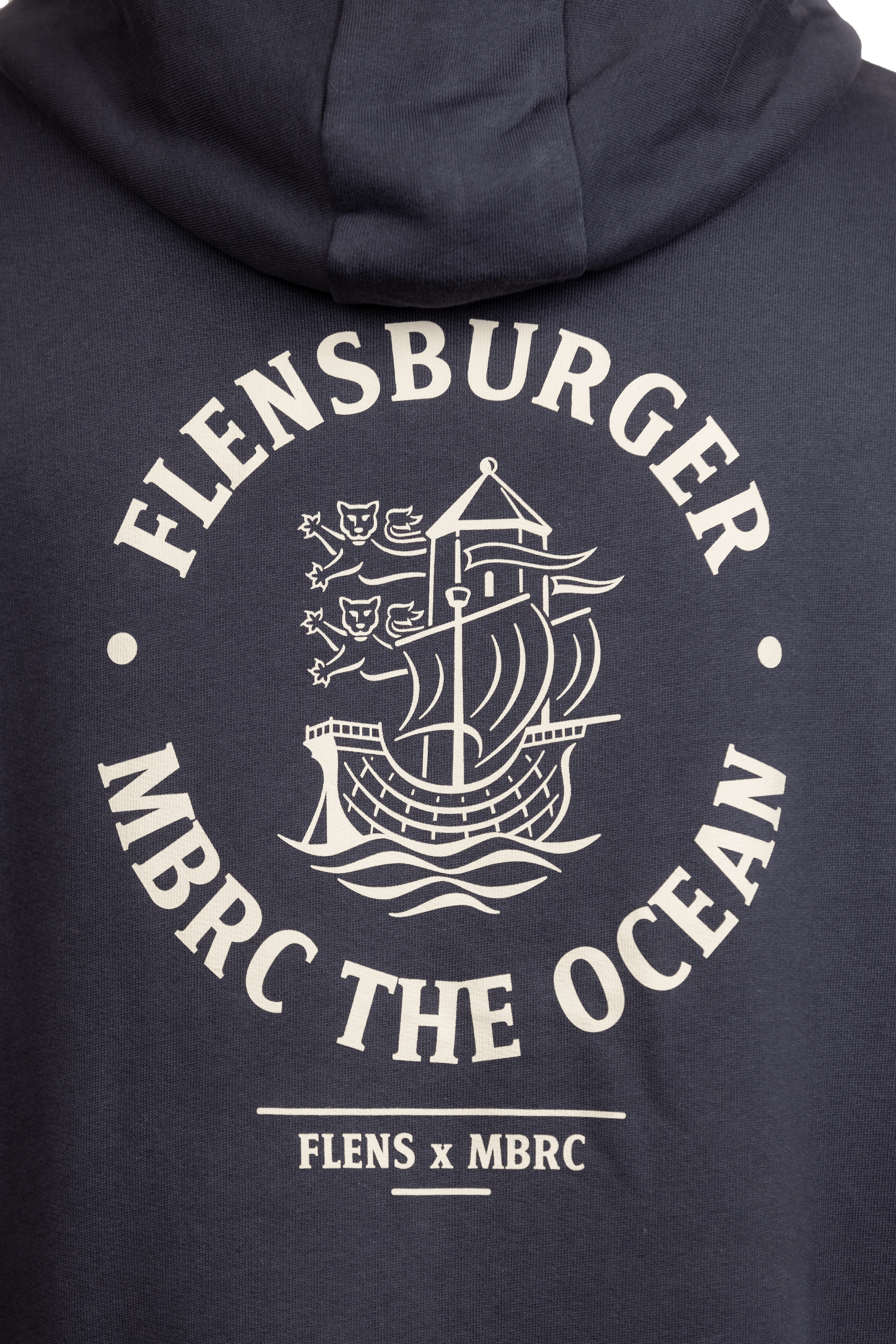 Detailansicht des Drucks auf der Rückseite eines anthrazitfarbenen Hoodies mit den Worten „FLENSBURGER MBRC THE OCEAN“ rund um ein stilisiertes Segelschiff und darunter „FLENS x MBRC“.