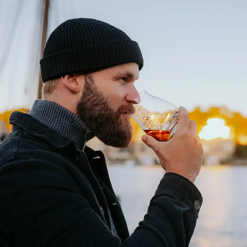 Ein bärtiger Mann trinkt genüsslich einen Schluck FLENS Whisky aus einem Tumbler-Glas im Sonnenuntergang am Hafen von Flensburg.