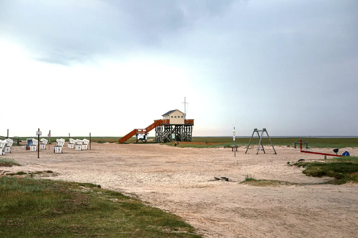 Nordsee-Strand von Sankt Peter-Ording mit Pfahlhaus auf Stelzen, Strandkörben und Spielplatz unter bewölktem Himmel.