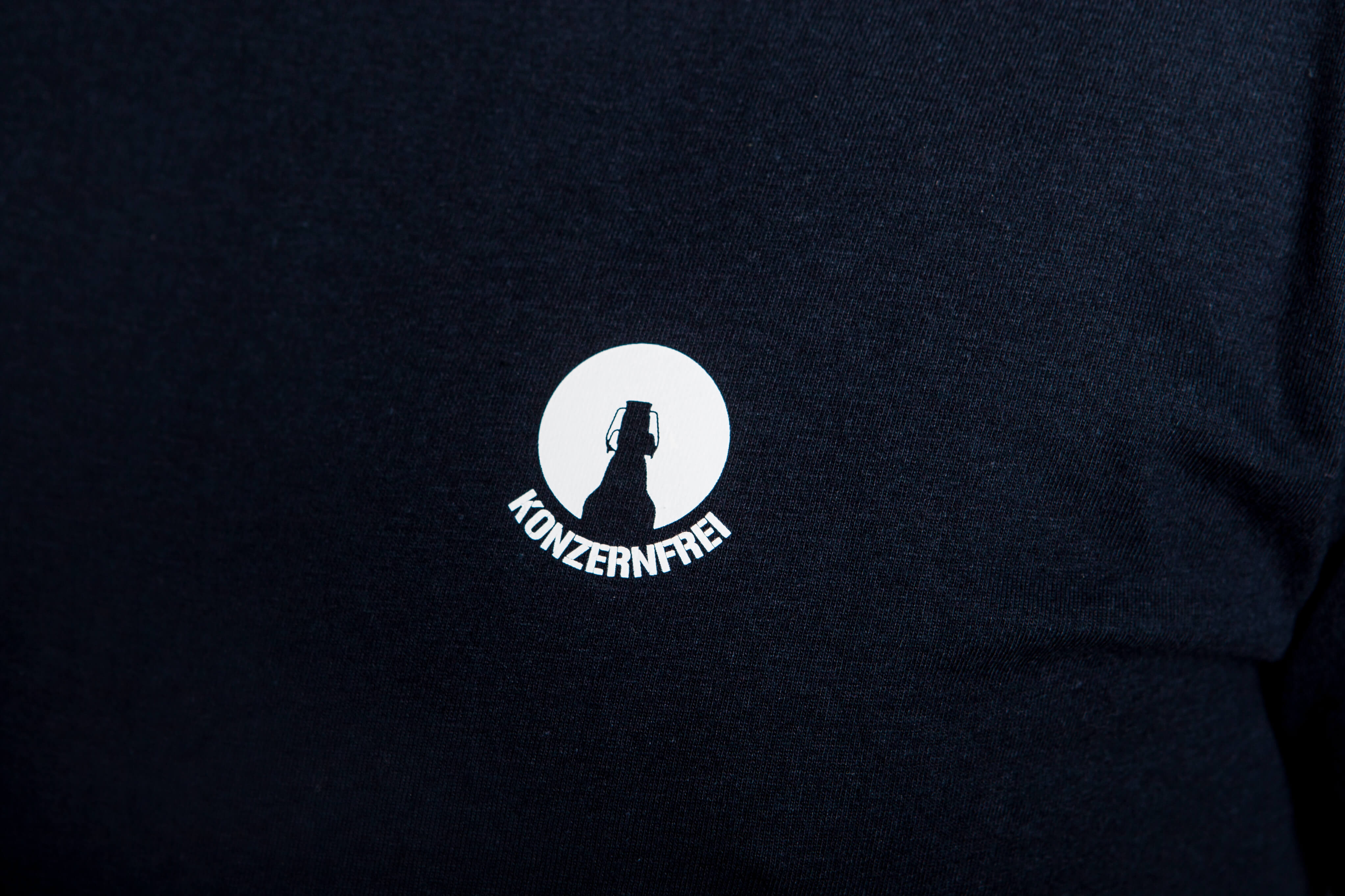 Weißer Aufdruck einer Flaschen-Silhouette in einem Kreis mit dem Text "KONZERNFREI" auf dunkelblauem Stoff eines T-Shirts.