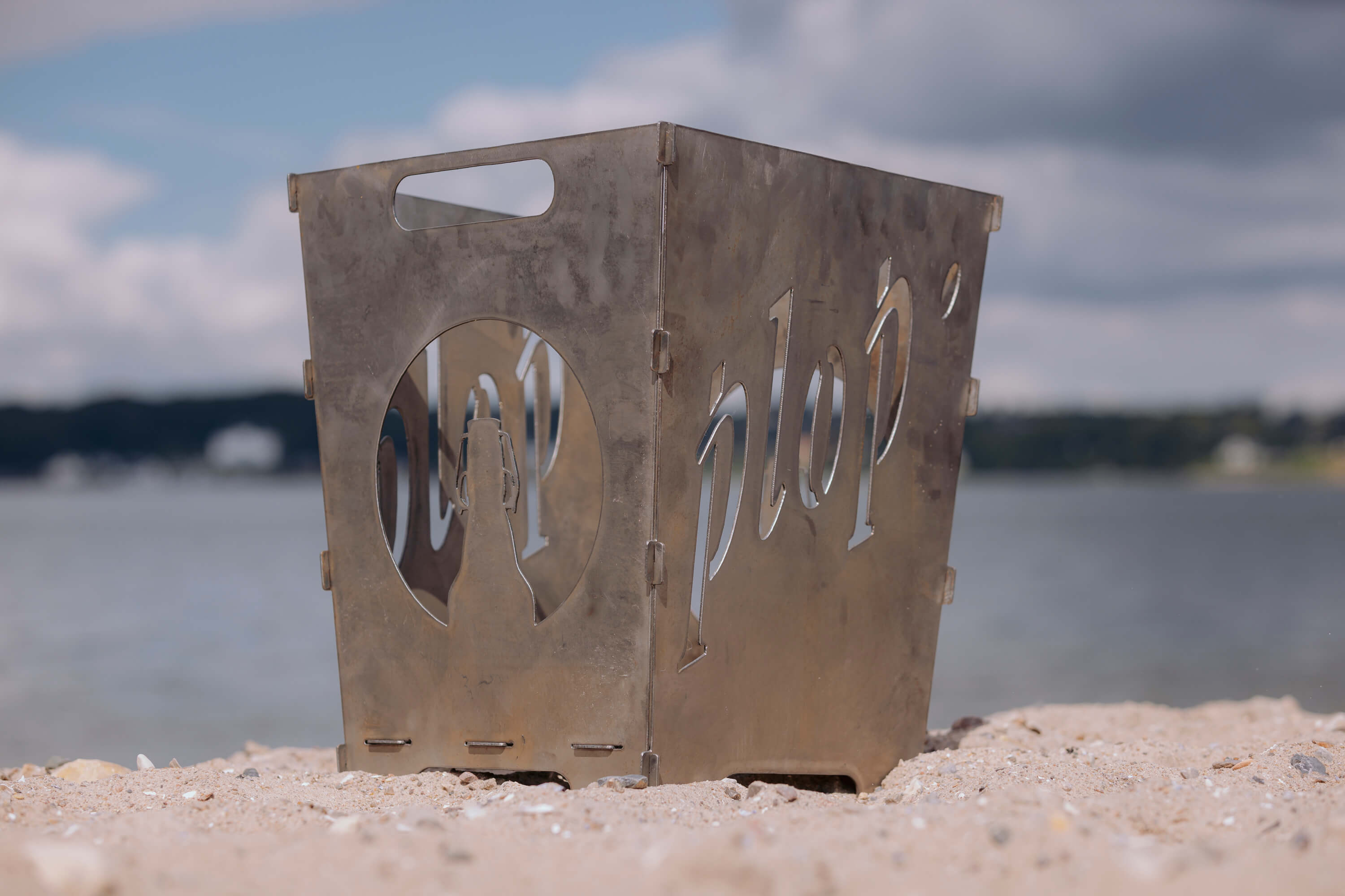 Metallener FLENS Feuerkorb mit ausgeschnittenem Schriftzug "plop'" und Flaschenmotiv steht auf Sand vor Wasser und bewölktem Himmel.