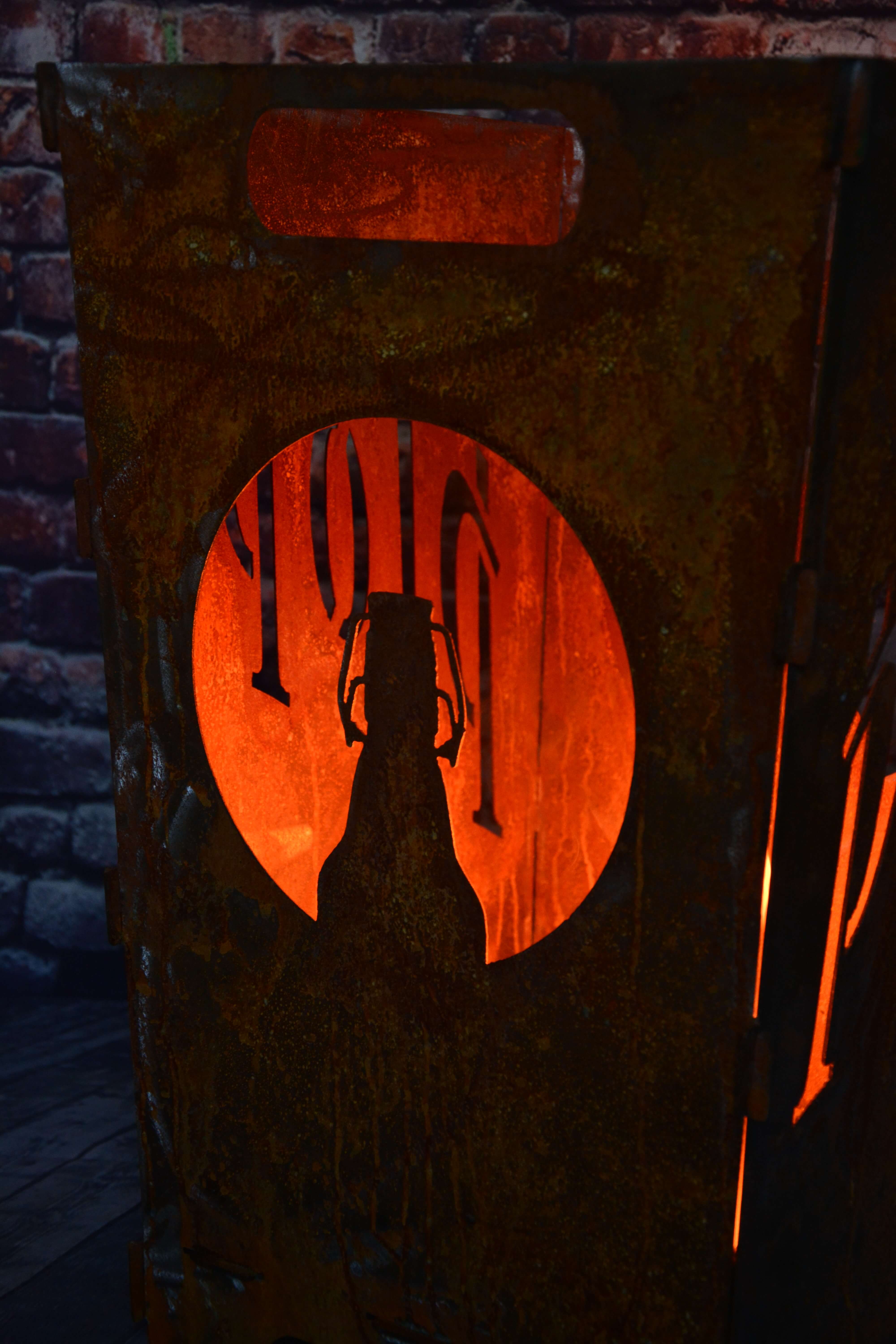 Feuerkorb in Rostoptik mit einem runden Ausschnitt, der die Silhouette einer Bügelflasche vor einem orangefarbenen Hintergrund zeigt. Im Hintergrund ist eine Wand aus roten Ziegelsteinen zu sehen.