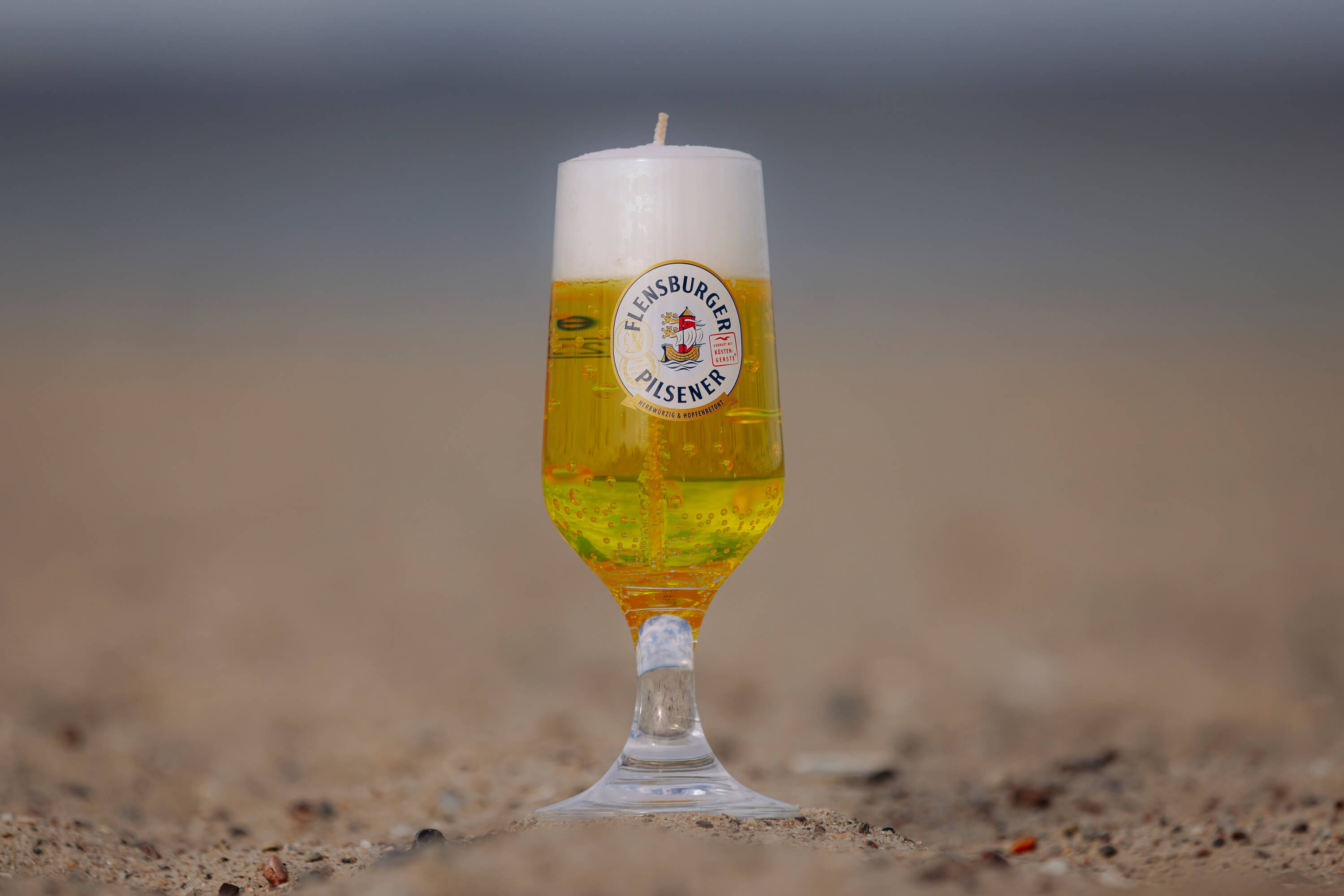 In einem 0,3 l Pokalglas ist eine Kerze, die goldenes Bier und Schaum darstellt. Auf dem Glas ist das Logo „Flensburger Pilsener“ zu sehen. Das Glas steht im Sand.