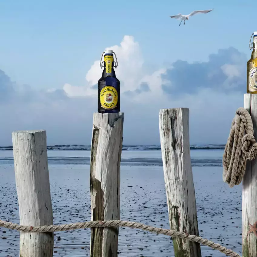 Vier Flensburger Flaschen stehen auf Holzstämmen am Meer.