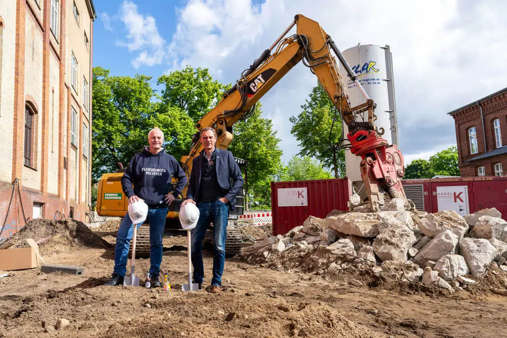 Die beiden Geschäftsführer der Brauerei stehen auf der Baustelle vorm Brauereigebäude vor einem großen Bagger.