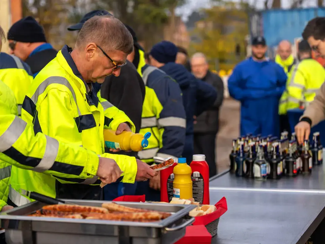 Arbeiter in Sicherheitskleidung servieren Grillwürstchen, im Hintergrund viele Flaschen mit alkoholfreiem FLENS.