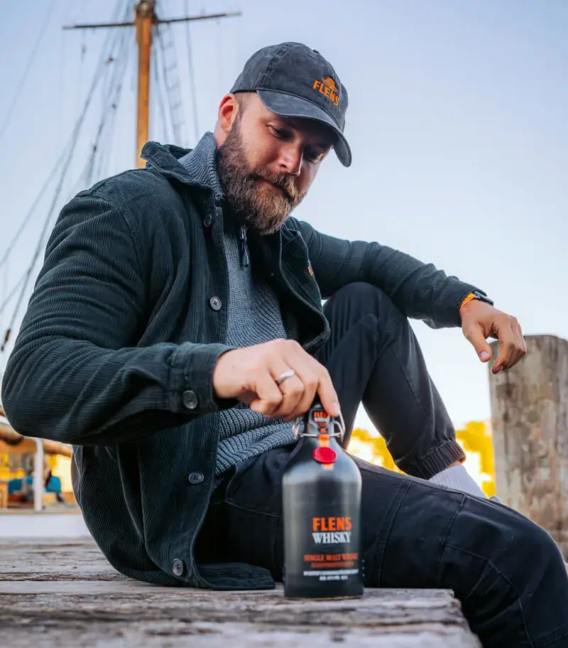 Mann im Whisky-Merch sitzt am Hafen mit der FLENS-Whisky-Flasche, der Schifffahrtsedition aus 2025.