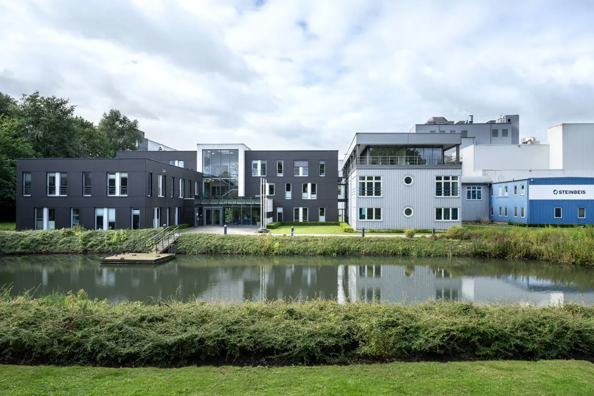 Modernes Büro- und Fabrikgebäude von Steinbeis an einem Teich mit Glasfassade, Grünanlage und ruhiger Wasserspiegelung.