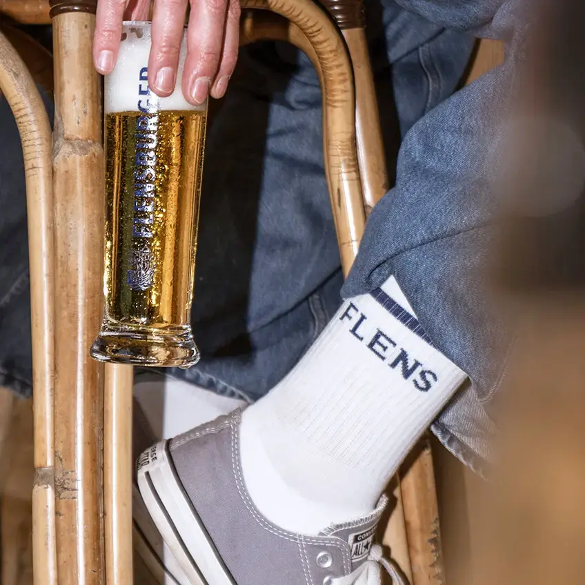 Hand hält Flensburger Pilsener im Stangenbierglas, Jeans, weiße FLENS Socken und graue Converse an Barhocker.